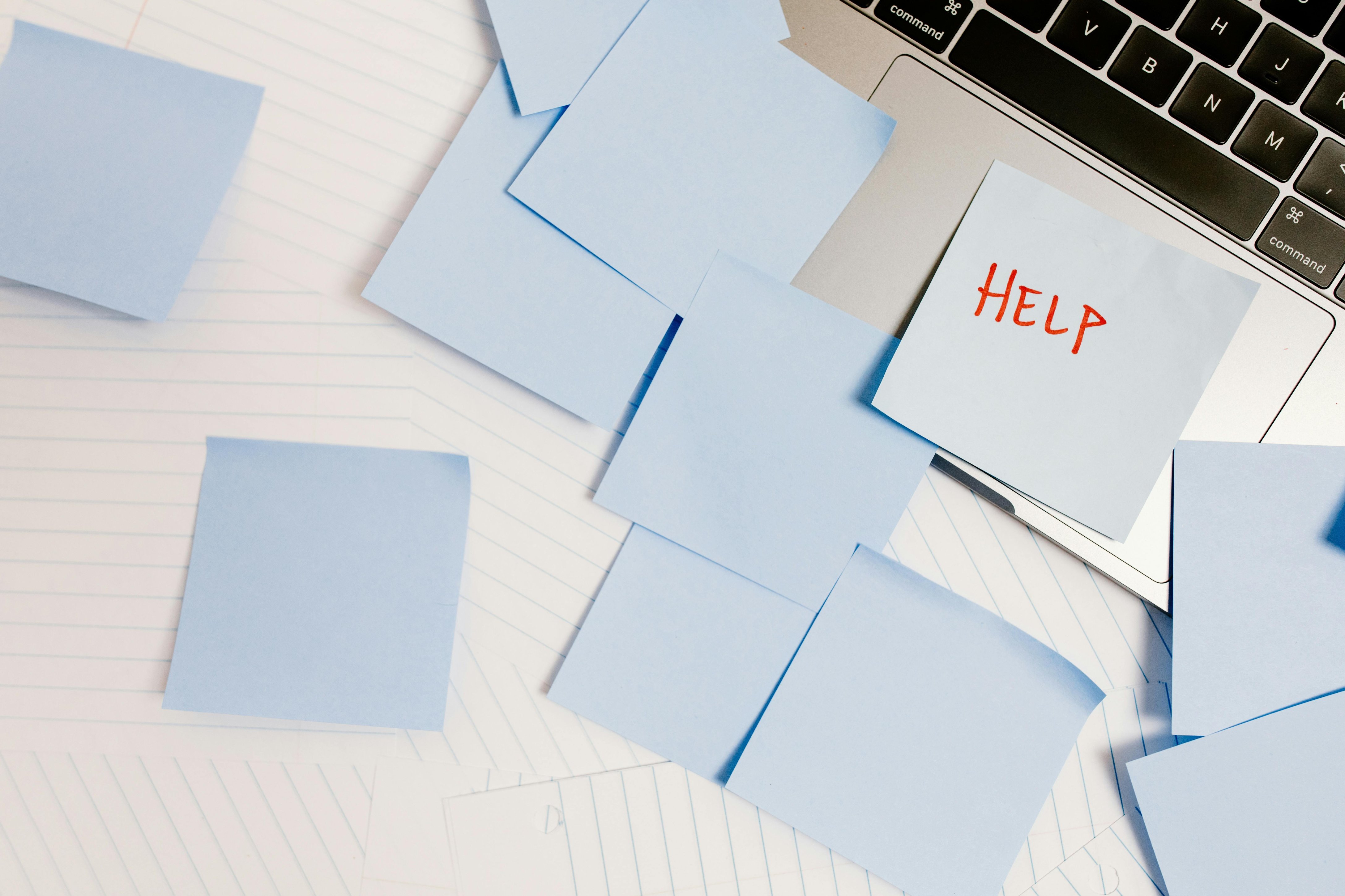 A cluttered desk with blue sticky notes scattered around a laptop, one note reading “HELP” in red letters.